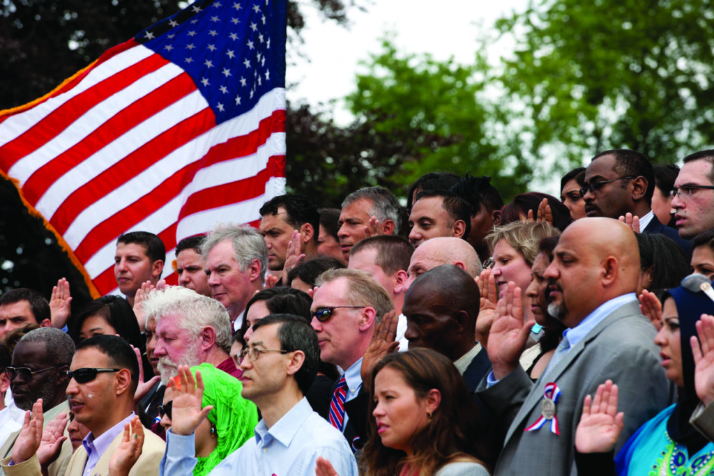Crowd of people raising their right hands and pledging at a naturalization ceremony at Thomas Jefferson's Monticello. An American flag waves in the background.